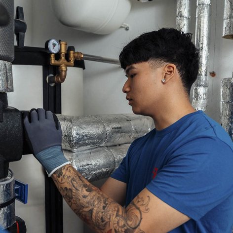 Ein junger Mann mit Tattoos arbeitet an einer Rohrleitung, während ein Kollege ihm aufmerksam zusieht.