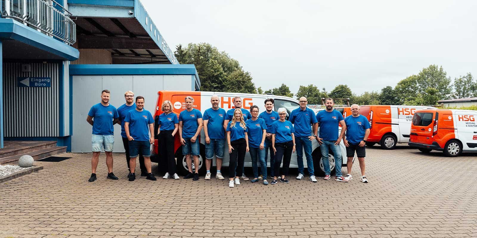 Eine Gruppe von zehn Personen in blauen T-Shirts steht vor einem Bürogebäude und mehreren orangefarbenen Lieferwagen.