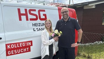 Eine junge Frau überreicht einem Mann mit Brille einen Blumenstrauß vor einem Firmenfahrzeug mit dem Logo HSG.