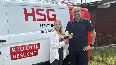 Freundliche Begegnung vor dem HSG-Fahrzeug im Freien Eine junge Frau überreicht einem Mann mit Brille einen Blumenstrauß vor einem Firmenfahrzeug mit dem Logo HSG.