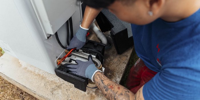 Eine Person mit tätowierten Armen arbeitet an einem Gerät, während sie mit einem Werkzeug in der Hand eine Reparatur durchführt.