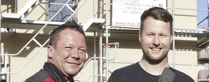 Zwei Männer lächeln vor einem Baugerüst, der eine trägt eine rote Jacke, der andere ein schwarzes Oberteil.