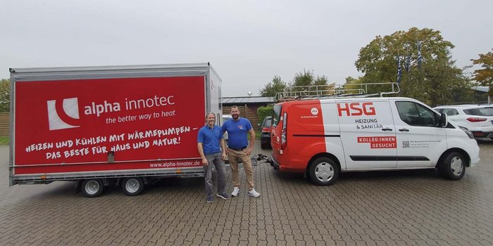 Zwei Männer stehen lächelnd vor einem roten Transporter mit dem Logo von alpha innotec und einem weißen HSG-Fahrzeug.