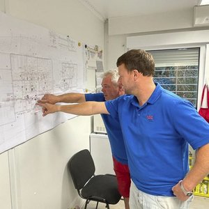 Zwei Männer in blauen T-Shirts betrachten einen großen Plan an der Wand und zeigen mit dem Finger auf verschiedene Punkte.
