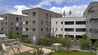 Moderne Wohngebäude umgeben von grünen Bäumen und einem Spielplatz mit Sandkasten und Klettergerüst im Vordergrund.