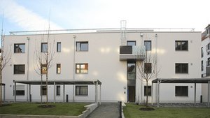 Ein modernes, helles Gebäude mit großen Fenstern und einem klaren, minimalistischen Design, umgeben von jungen Bäumen.