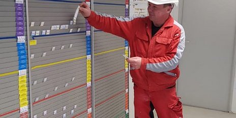 Ein Arbeiter in roter Arbeitskleidung und Helm steht vor einer Wand mit farbigen Schildern und notiert Informationen.