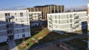 Moderne Wohngebäude umgeben von einem gepflegten Garten mit Bäumen und Sitzgelegenheiten, unter einem klaren blauen Himmel.