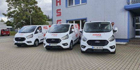 Drei weiße Lieferwagen mit HSG-Logo stehen vor einem modernen Gebäude, während zwei Personen lächeln und winken.