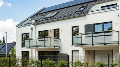 Ein modernes, zweigeschossiges Wohnhaus mit großen Fenstern und einem Balkon, umgeben von grünen Pflanzen und Bäumen.