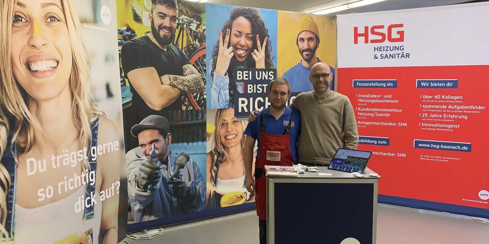 Zwei Männer stehen an einem Messestand, umgeben von bunten Plakaten mit lächelnden Gesichtern und Handwerksmotiven.