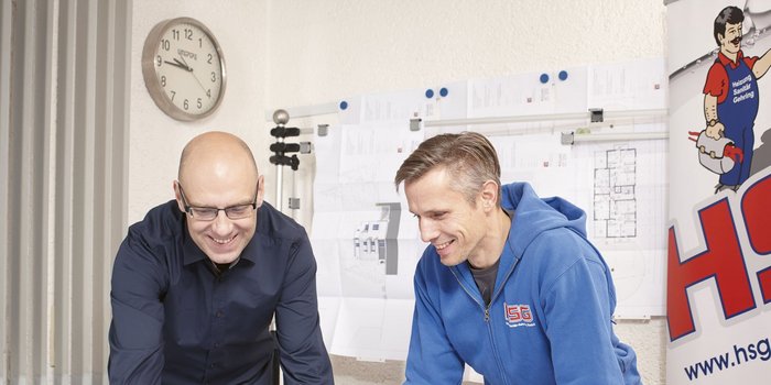 Zwei Männer in einem Büro betrachten gemeinsam Pläne, während eine Wanduhr im Hintergrund die Zeit anzeigt.