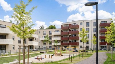 Eine moderne Wohnanlage mit mehreren Etagen, umgeben von einem gepflegten Garten und einem Spielplatz für Kinder.