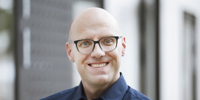 Ein lächelnder Mann mit Glatze und Brille trägt ein dunkelblaues Hemd, vor einem unscharfen Hintergrund.