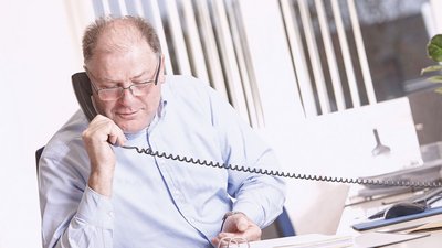 Ein älterer Mann mit Brille spricht konzentriert am Telefon, während er an einem Schreibtisch mit Unterlagen sitzt.