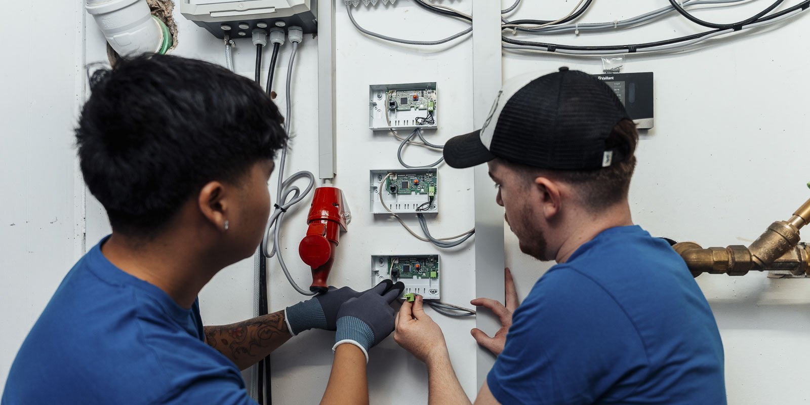Zwei Techniker in blauen T-Shirts arbeiten konzentriert an elektrischen Steuerkästen an einer Wand.