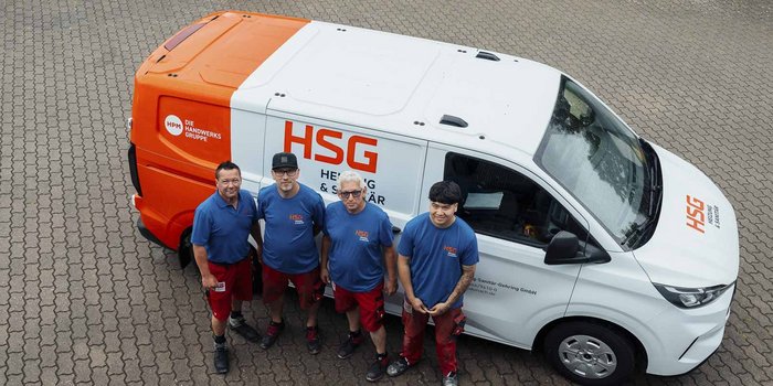 Vier Handwerker stehen lächelnd vor einem orange-weißen Lieferwagen mit dem Logo von HSG auf einem gepflasterten Boden.