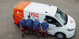 Vier Handwerker stehen lächelnd vor einem orange-weißen Lieferwagen mit dem Logo von HSG auf einem gepflasterten Boden.