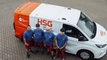 Vier Handwerker stehen lächelnd vor einem orange-weißen Lieferwagen mit dem Logo von HSG auf einem gepflasterten Boden.
