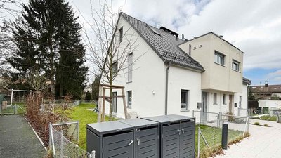 Die Fassade eines hellen Hauses mit einem schrägen Dach, umgeben von einem eingezäunten Garten und einem Spielplatz.
