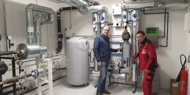 Zwei Männer stehen in einem technischen Raum, umgeben von Rohren und Maschinen, während sie an einem Gerät arbeiten.