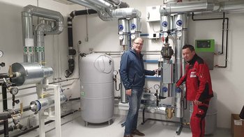 Zwei Männer stehen in einem technischen Raum, umgeben von Rohren und Maschinen, während sie an einem Gerät arbeiten.