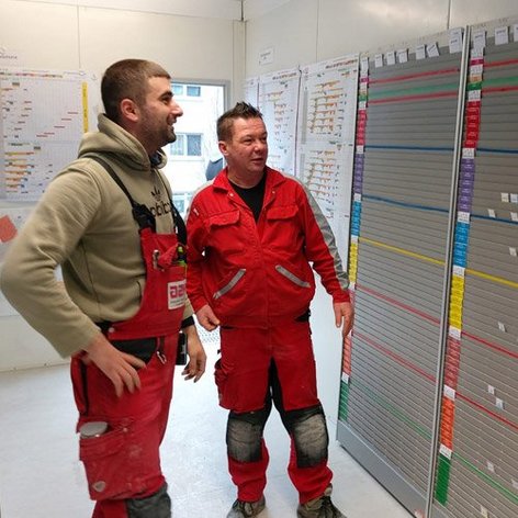 In einem hellen Raum unterhalten sich zwei Arbeiter in roten Overalls, während sie eine farbige Wandtafel mit Plänen betrachten.