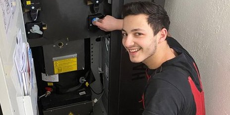 Ein junger Techniker in roter Arbeitskleidung lächelt, während er an einem technischen Gerät arbeitet.