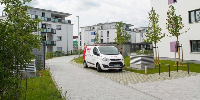 Ein Lieferwagen steht auf einem gepflasterten Weg in einer modernen Wohnanlage mit grünen Bäumen im Hintergrund.